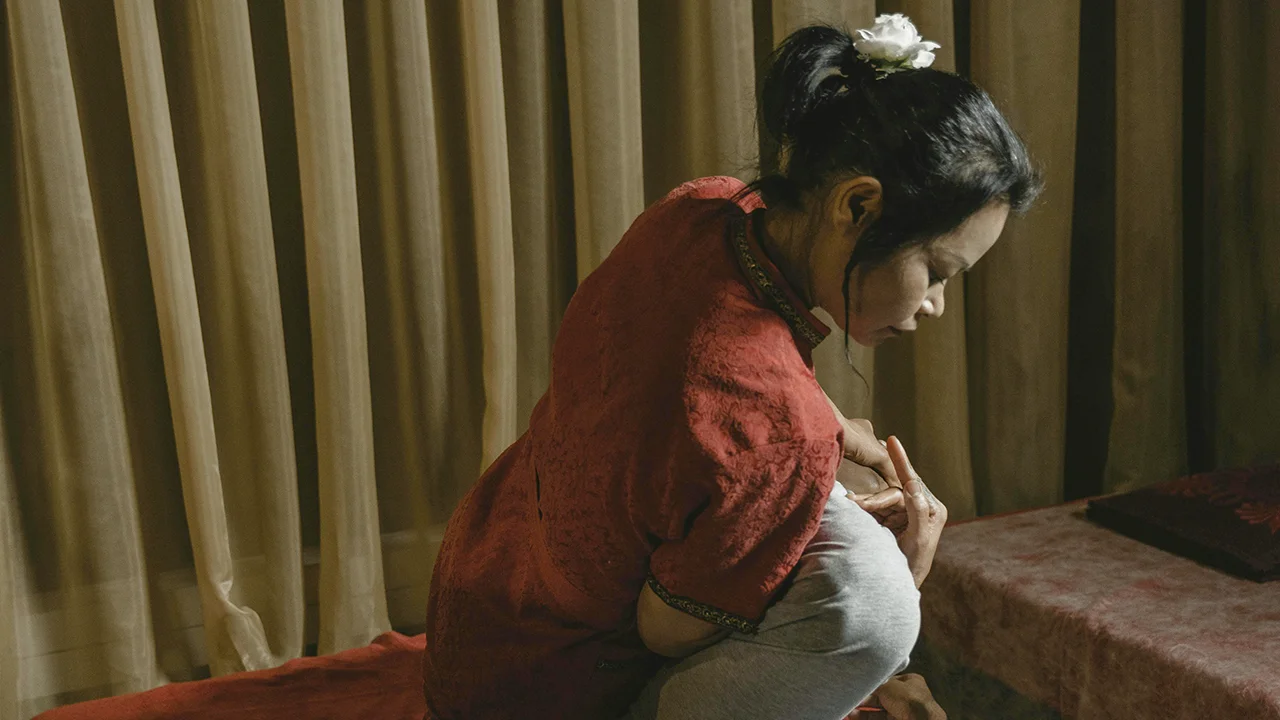Une praticienne en tenue rouge avec une fleur blanche dans les cheveux réalise un massage assis sur une personne allongée, dans une pièce avec rideaux beiges et une table de massage recouverte de tissu rouge.