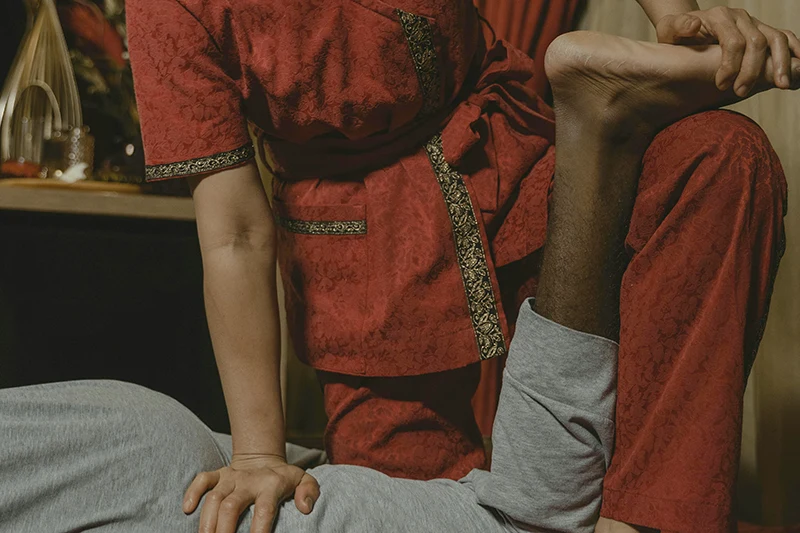 Un masseur en tenue rouge traditionnelle effectue un étirement de jambe à un client allongé portant un t-shirt gris lors d'un massage en apesanteur.