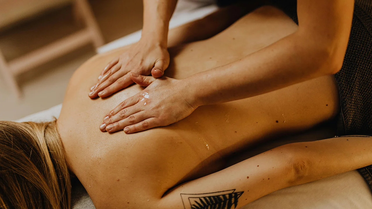 Une personne allongée sur le ventre reçoit un massage du dos par une autre personne appliquant de l'huile, avec un tatouage visible sur l'avant-bras.