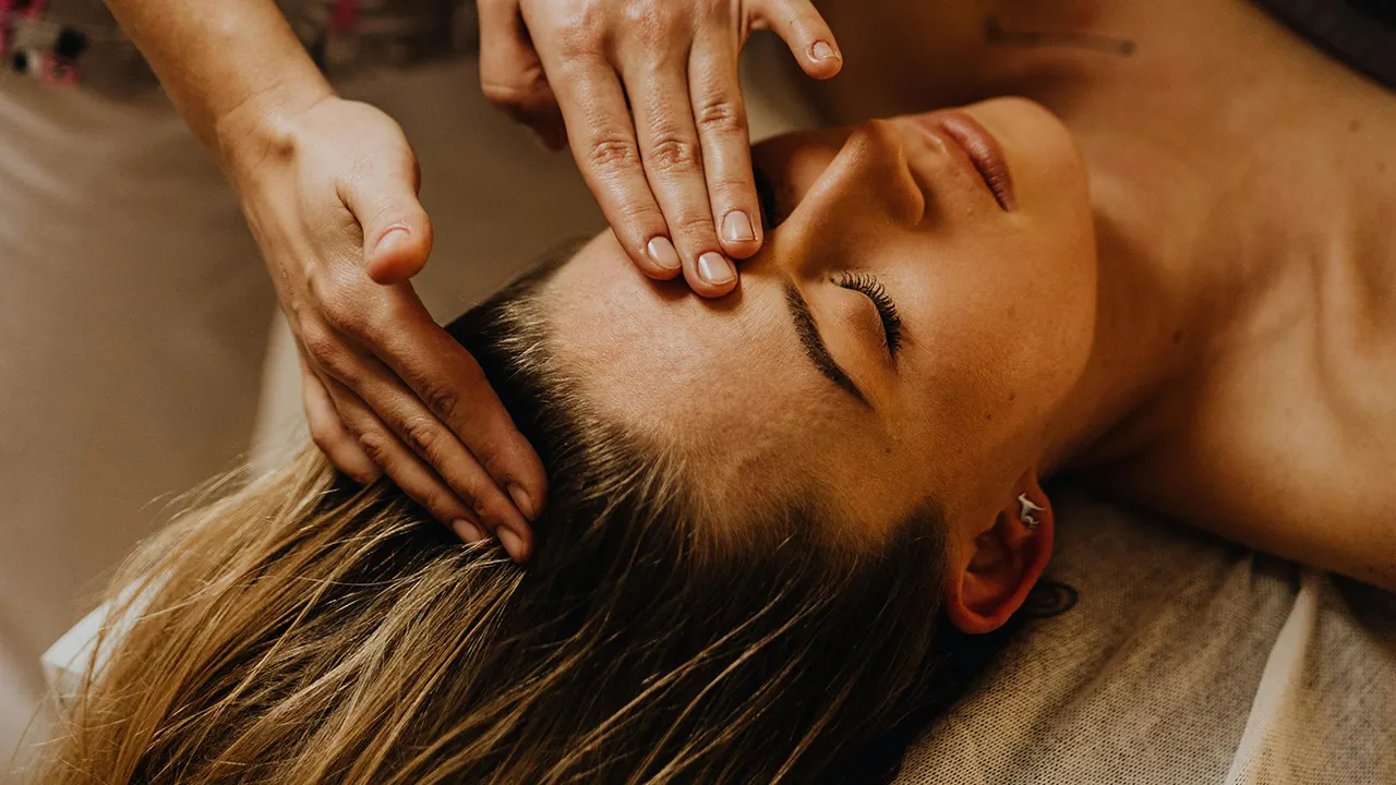 Une personne recevant un massage du visage avec les mains appliquées sur le front, yeux fermés, cheveux détachés, en position allongée.