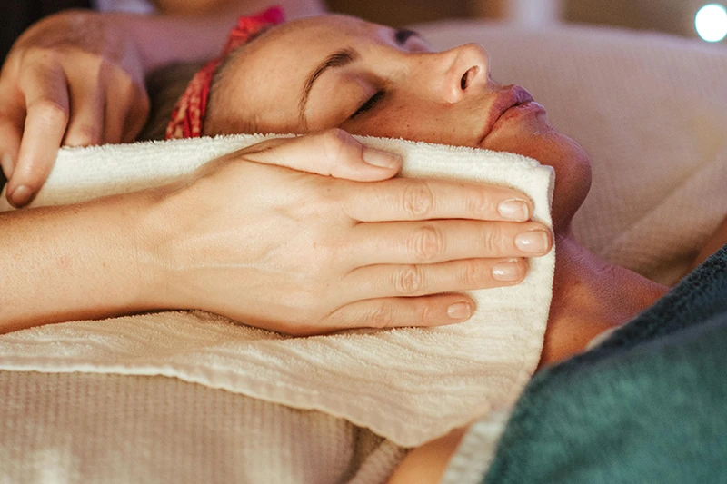 Une personne allongée reçoit un massage du visage avec une serviette blanche posée sur la joue, les yeux fermés, dans un cadre relaxant.