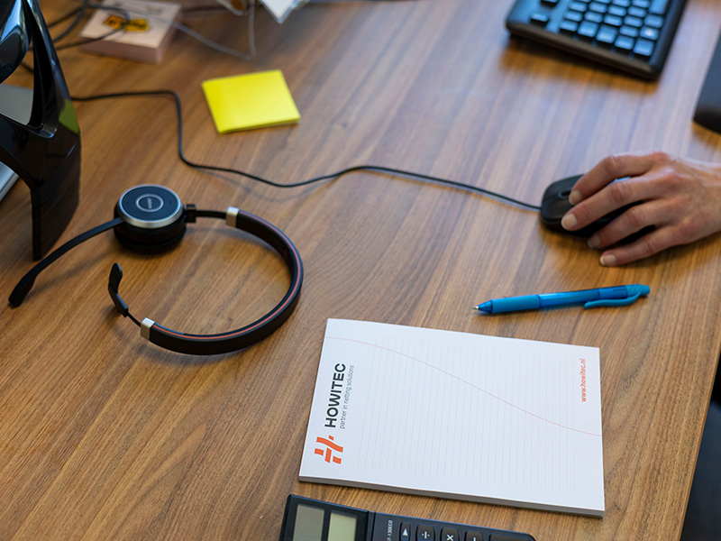 Bureaublad met een headset, computermuis, rekenmachine, pen, notitieblok en sticky note; een hand bedient de muis.