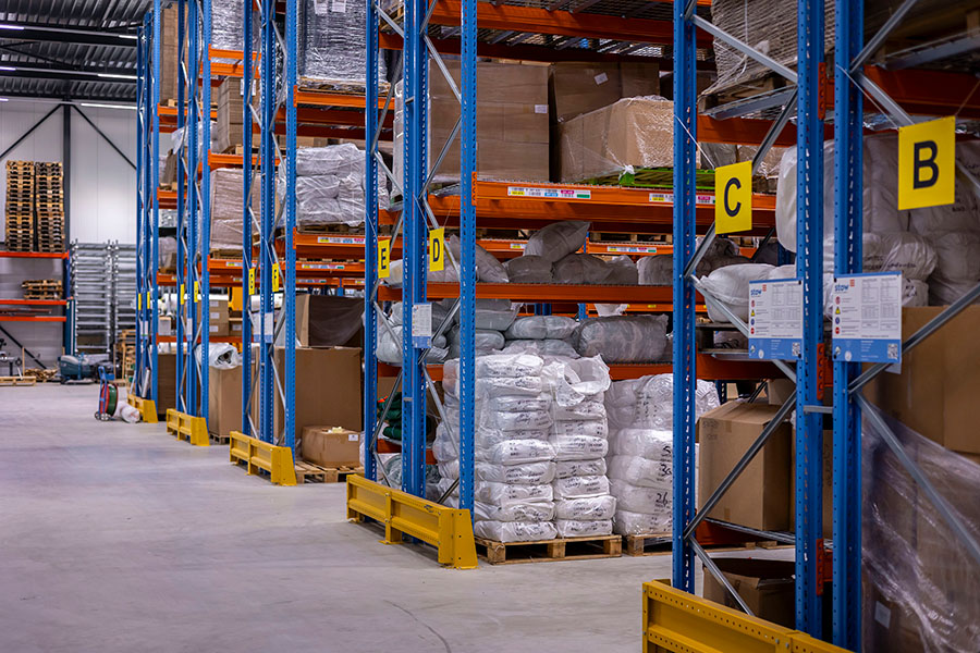 Magazijn met hoge stellingen vol pallets en verpakte goederen, voorzien van gele vaklabels en blauwe staanders.