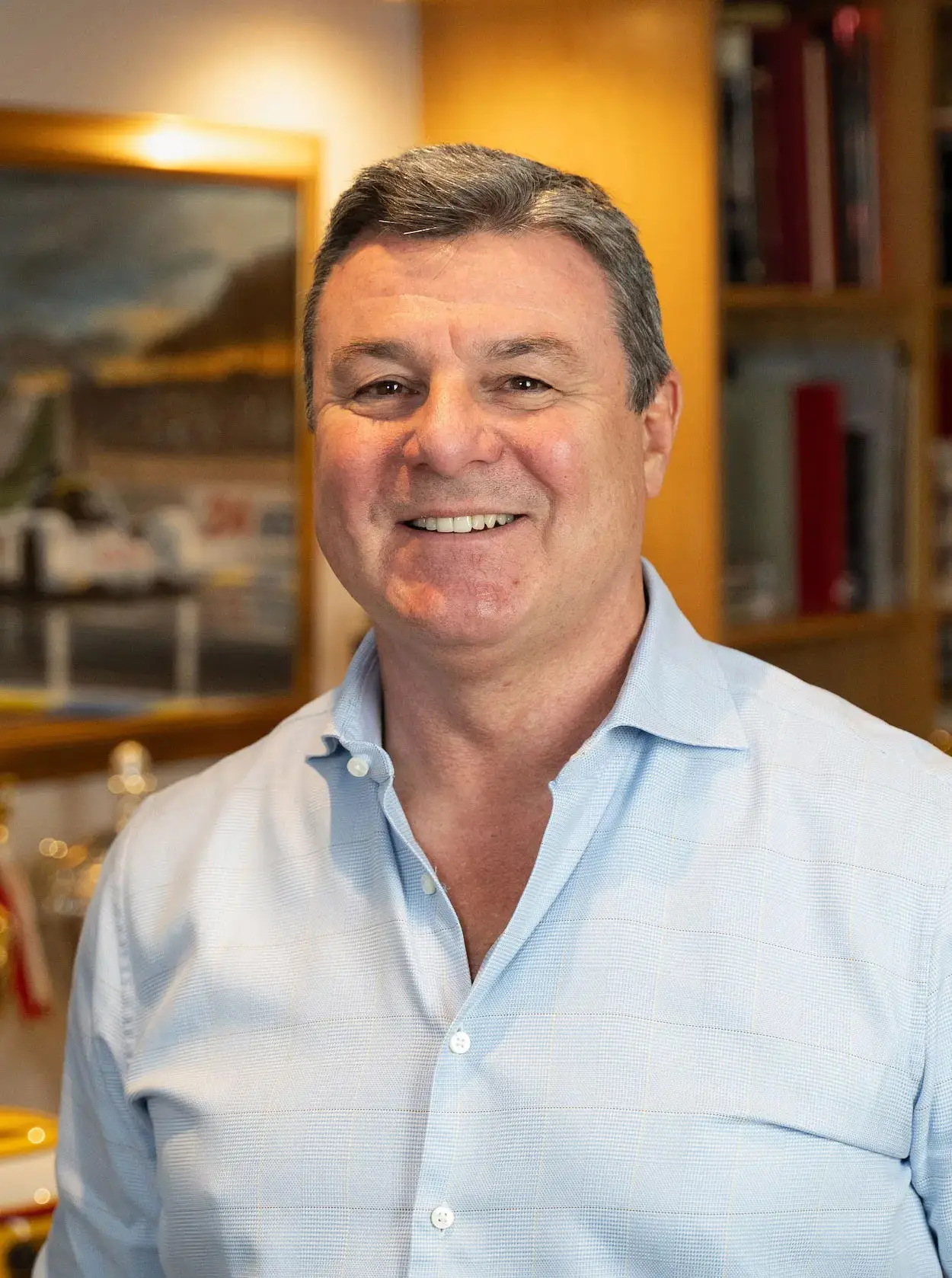 Smiling man wearing a light blue shirt standing indoors with a blurred background of bookshelves and framed picture.