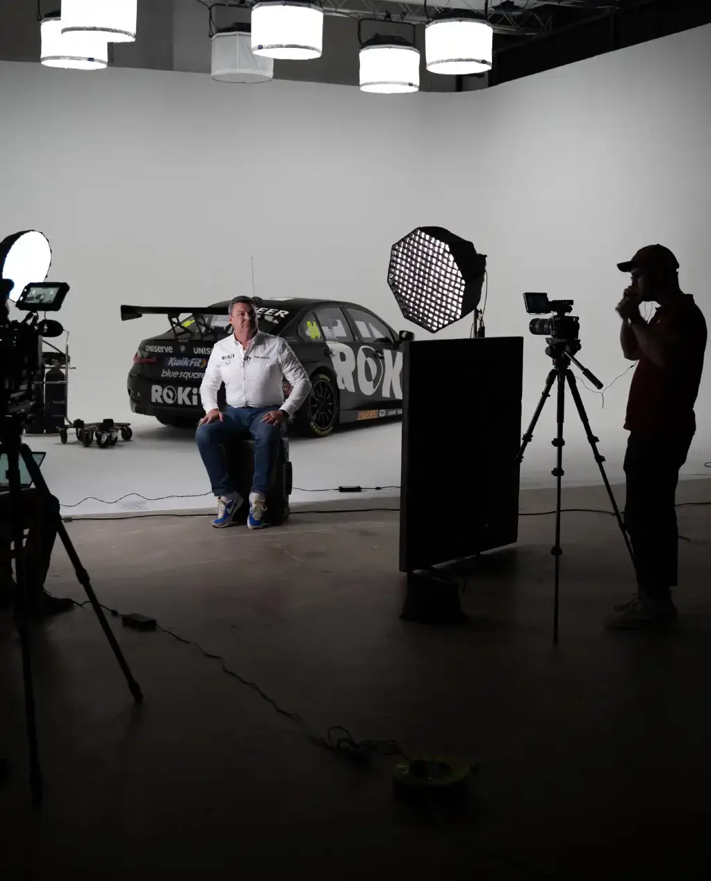 Young man wearing glasses and a black racing jacket sitting on the hood of a white and red Howdens sponsored race car outside a Howdens store.