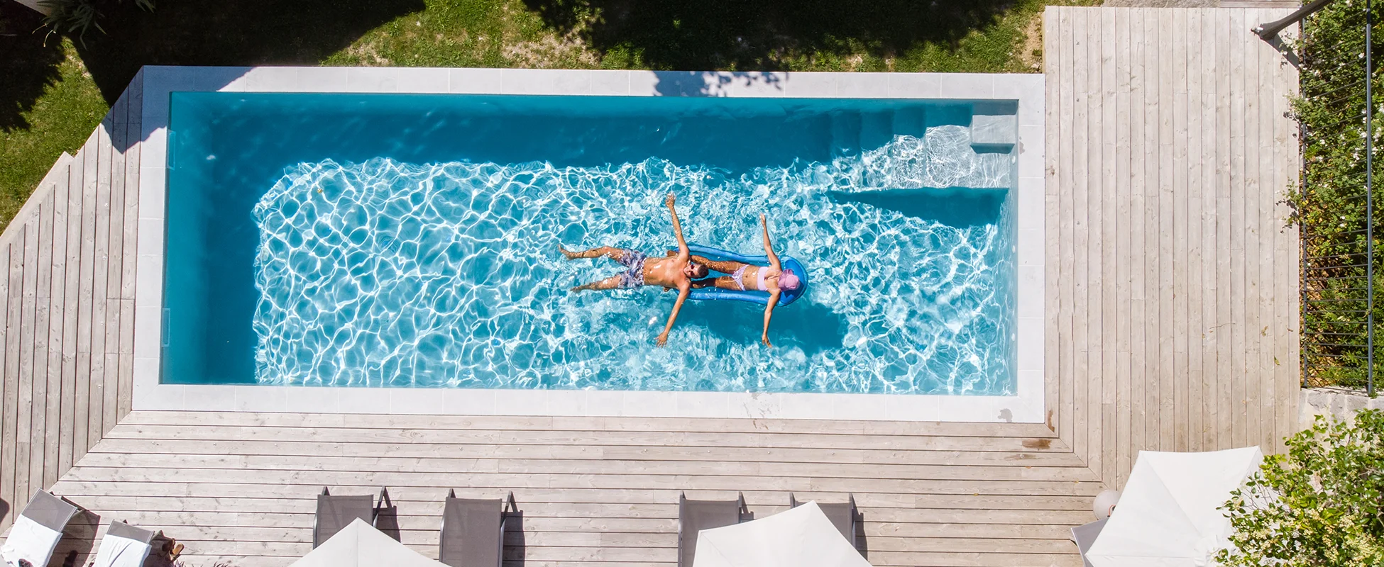 Overhead view of a man and a woman relaxing on a blue inflatable raft floating in a rectangular swimming pool surrounded by wooden deck and lounge chairs.