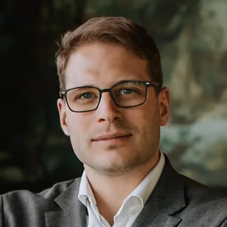 Tim Kantaut with glasses wearing a gray suit jacket and white shirt, neutral background.