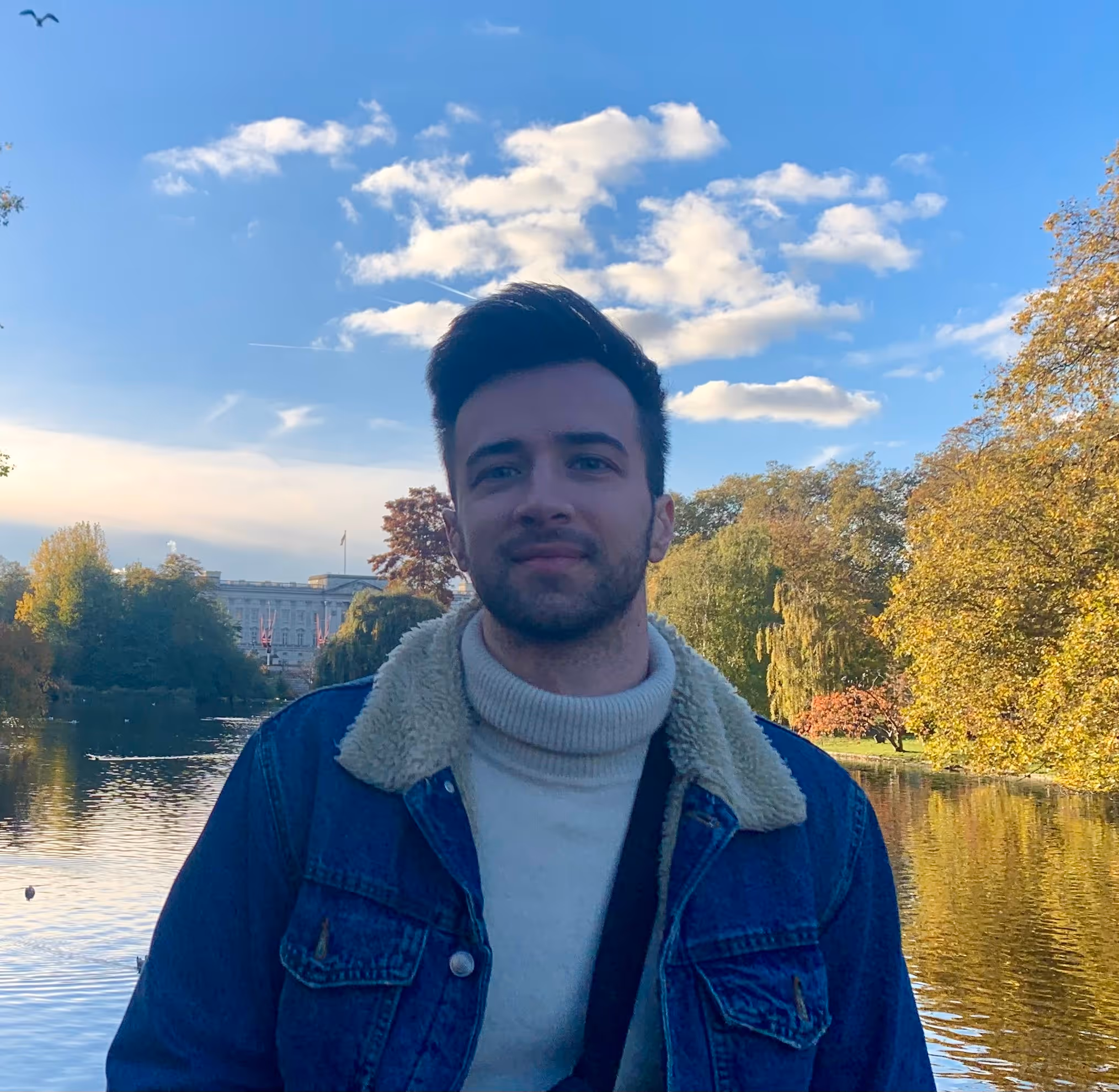 Young man wearing a denim jacket with a shearling collar and a white turtleneck sweater standing in front of a lake with autumn trees and a partly cloudy sky.