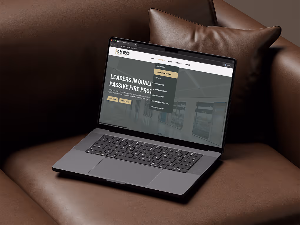 Open laptop on a brown leather sofa displaying a website for Kyro Fire Protection with a menu highlighting fire stopping translucent coatings.