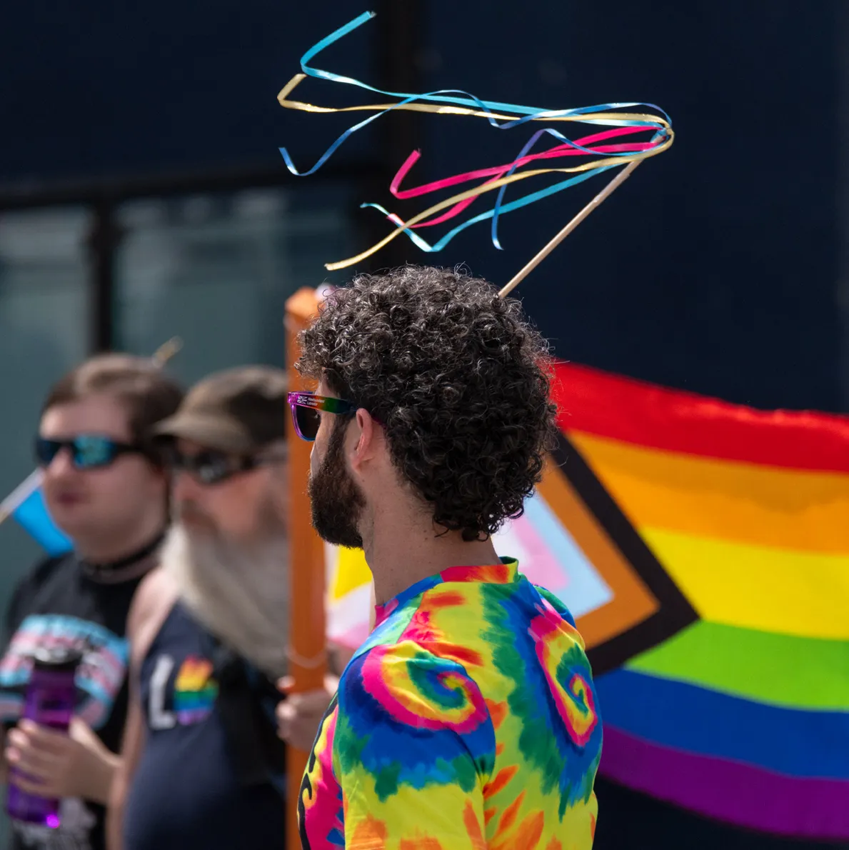 A person back on wearing a neon swirled t-shirt waving a party streamer in the air. There is a group of people holding a larger pride flag in the background.