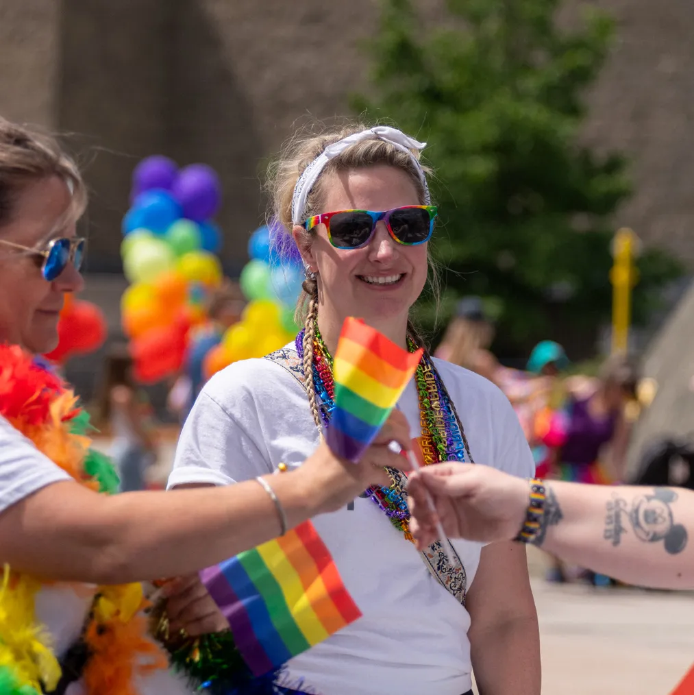 A person wearing rainbow sunglasses is smiling and passing a small pride flag to someone wearing a rainbow bracelet.