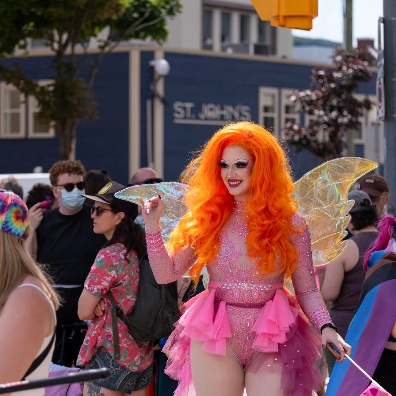 Drag Queen dressed in bright orange wig, pink detailed costume with fairy wings, smiling and holding a small pride flag at Pride.