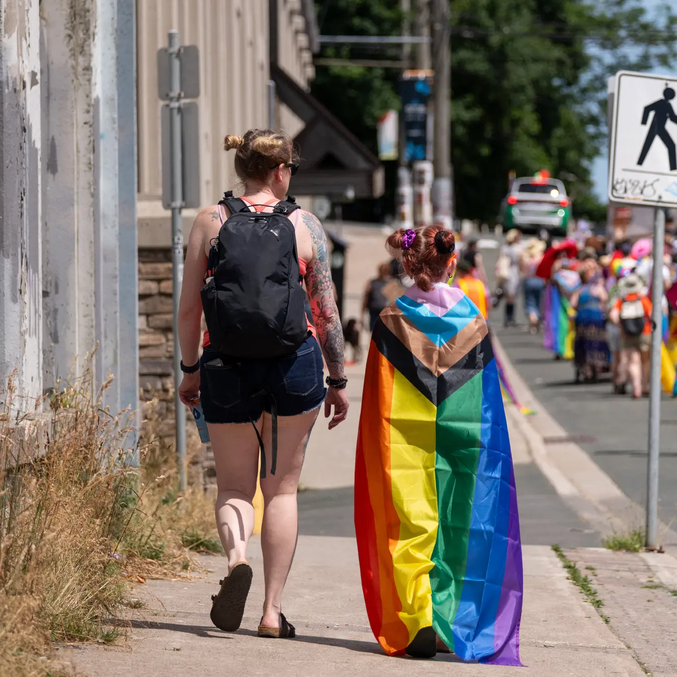 A mother and daughter are walking down the sidewalk looking at the passing parade. The little girl is wearing a pride flag as a cape.