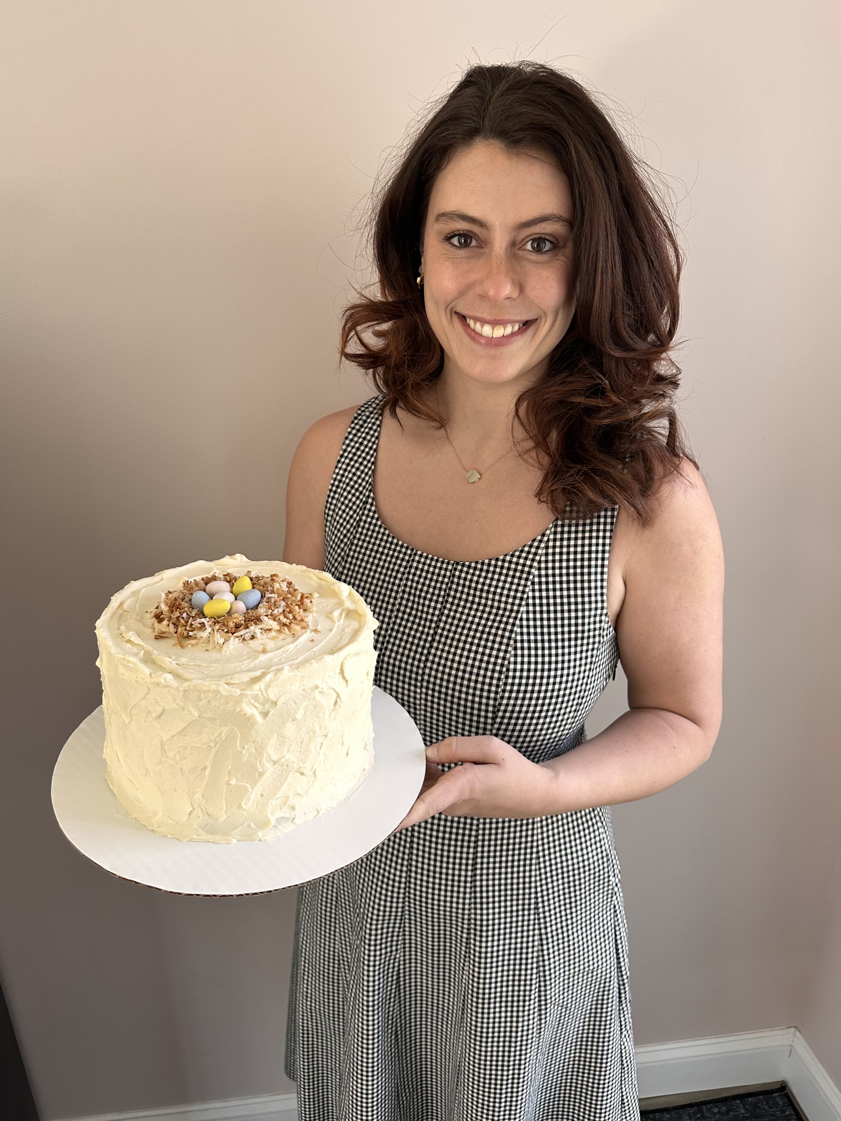 Smiling woman in black and white checkered dress holding a round white frosted cake decorated with toasted coconut and colorful candy eggs on top.