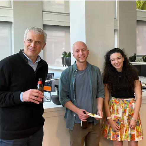 Three people smiling indoors, two men holding drinks and a woman wearing a colorful skirt.