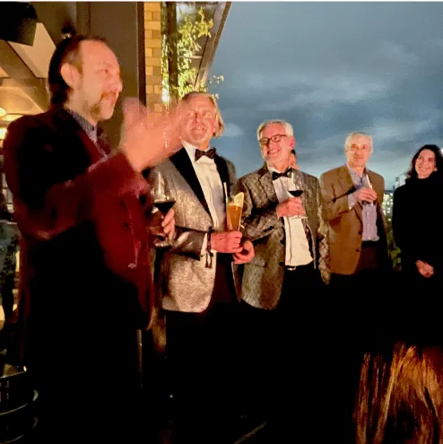 Group of people dressed formally holding drinks, socializing outdoors under a cloudy sky at dusk.