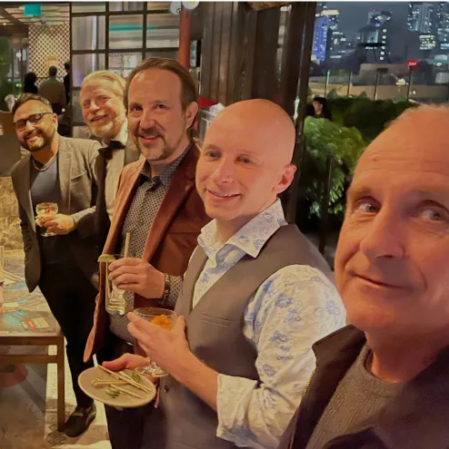 Five men in dressy casual attire smiling and holding drinks and snacks at a nighttime social event.