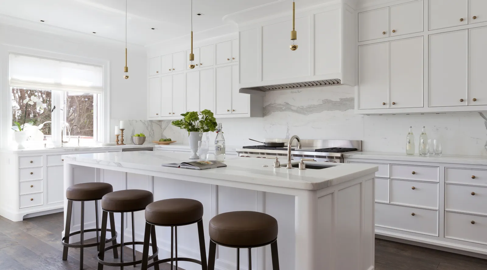 Bright white kitchen with marble countertops, four brown stools, pendant lights, and stainless stove.