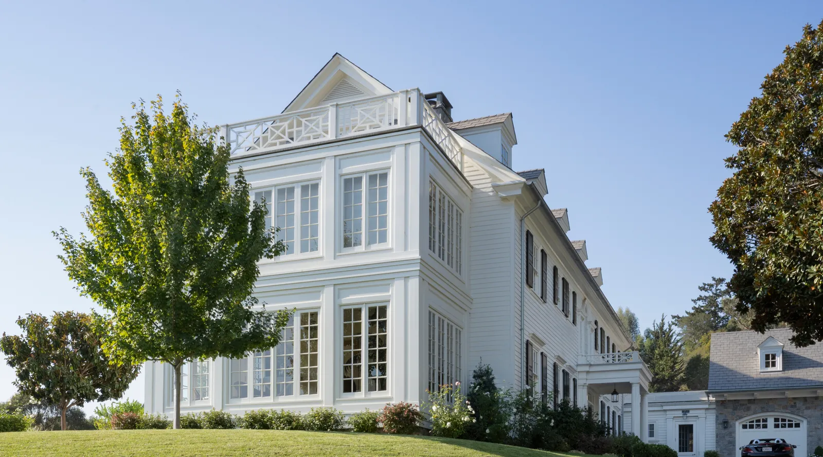 Large white multi-story house with numerous windows, a balcony, and trees on a grassy hill.