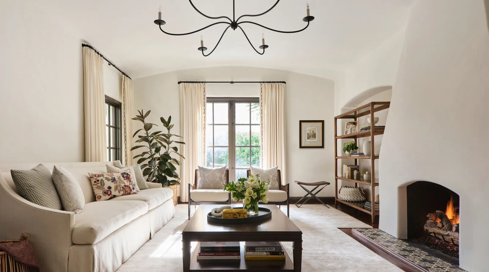 Bright living room with white sofa, two armchairs, wooden coffee table, bookshelf, and lit fireplace.