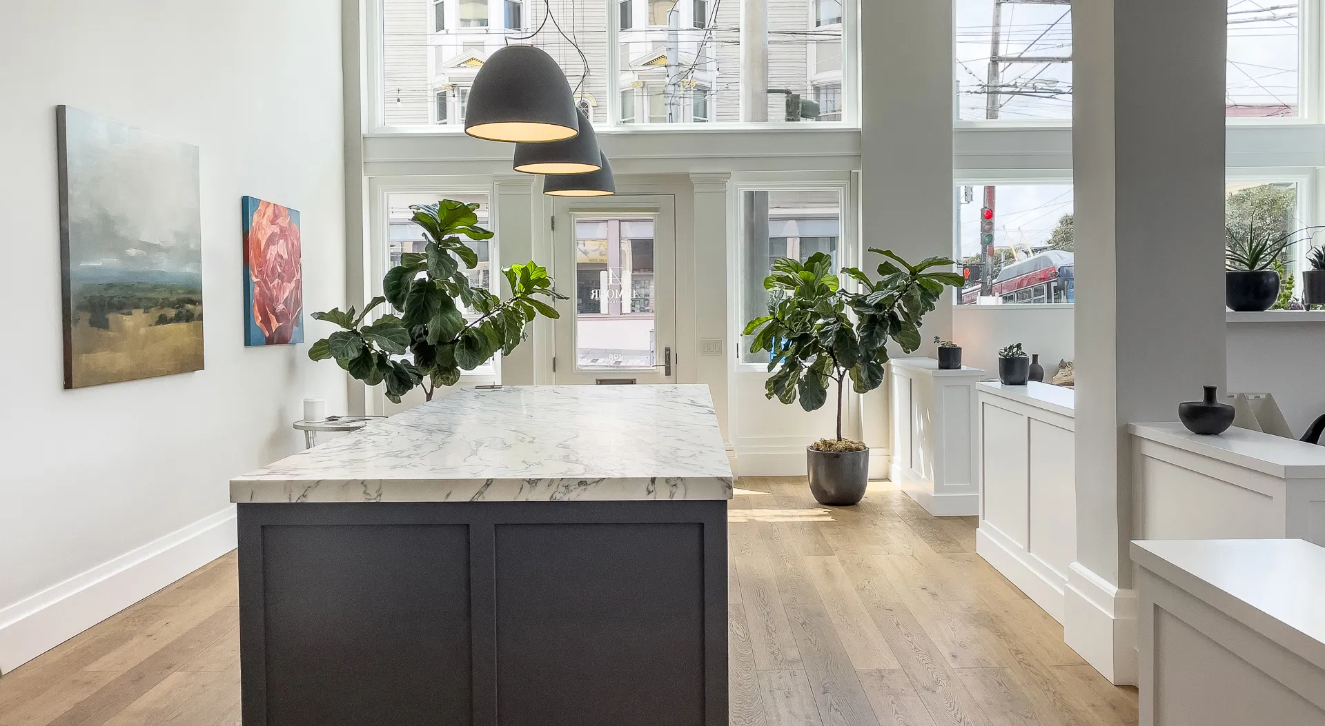 Bright modern office with marble island, hanging lamps, two large plants, artwork, and wooden floor.