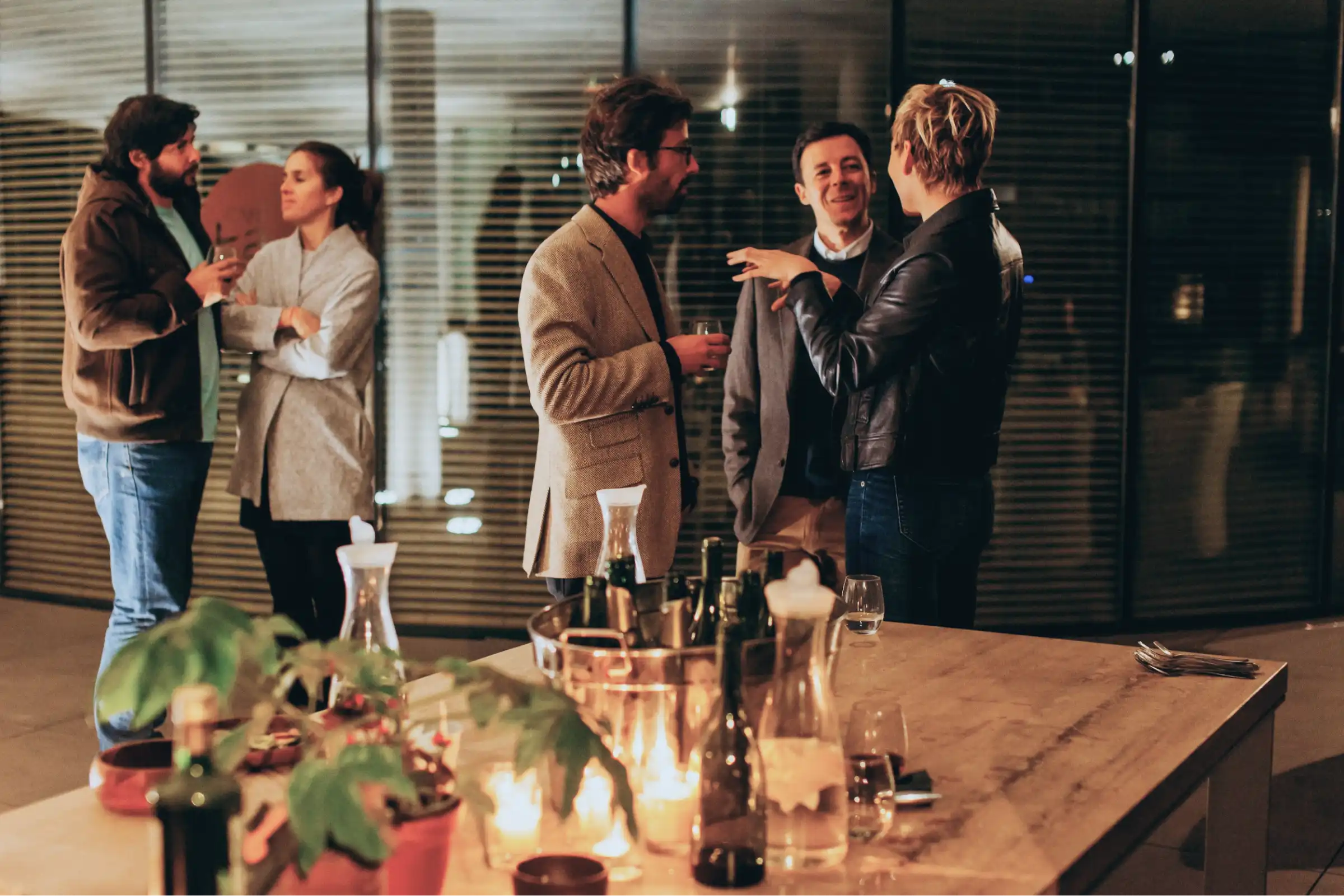 Two groups of people, mingling at an event, a table with drinks is in the foreground.  
