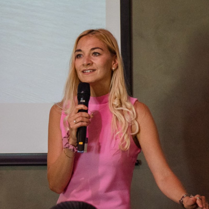 Blonde woman in a pink sleeveless top holding a microphone and speaking in front of a screen.