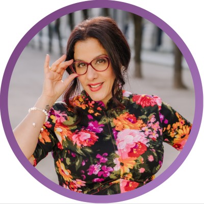 Woman with dark hair and glasses wearing a colorful floral dress, touching her glasses and smiling with a blurred outdoor background.