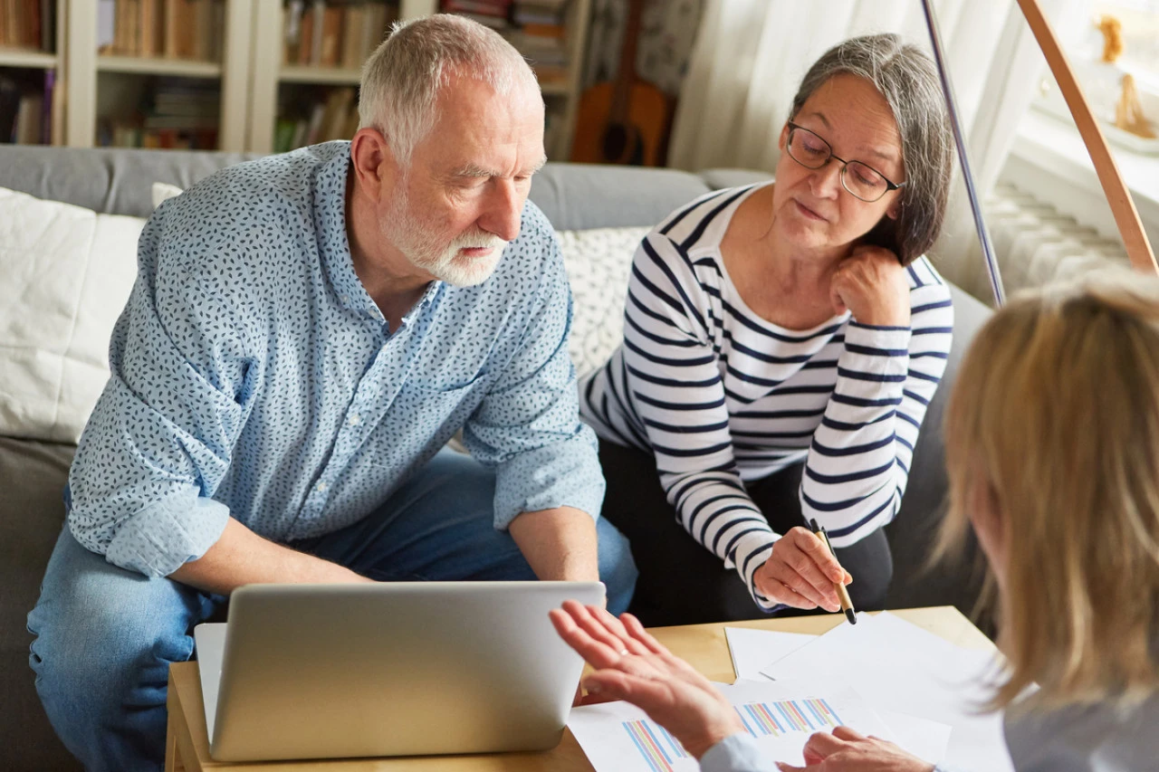 Un couple senior discute avec une ergothérapeute en regardant les informations nécessaires à la création d'un dossier MaPrimeAdapt' sur un ordinateur portable sur une table basse.