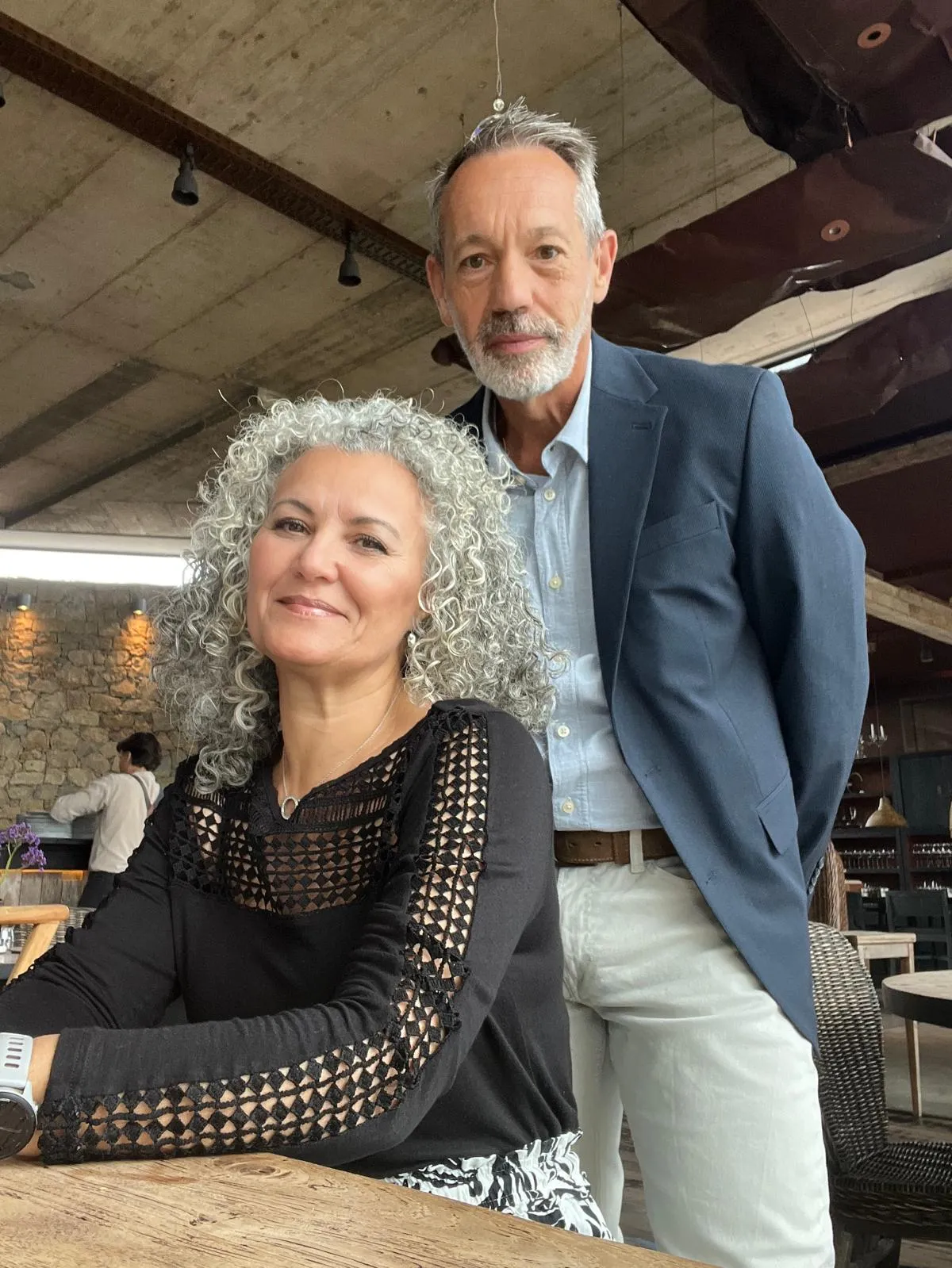 Smiling middle-aged woman with curly gray hair wearing a black top with lace sleeves seated at a wooden table, with a standing man behind her wearing a blue blazer, light blue shirt, white pants, and a brown belt, inside a restaurant with stone walls and industrial ceiling.