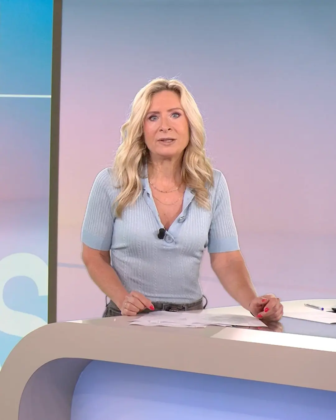 Female RTL Nieuws presenter with blonde hair in a light blue short-sleeve top standing behind a news desk with papers.