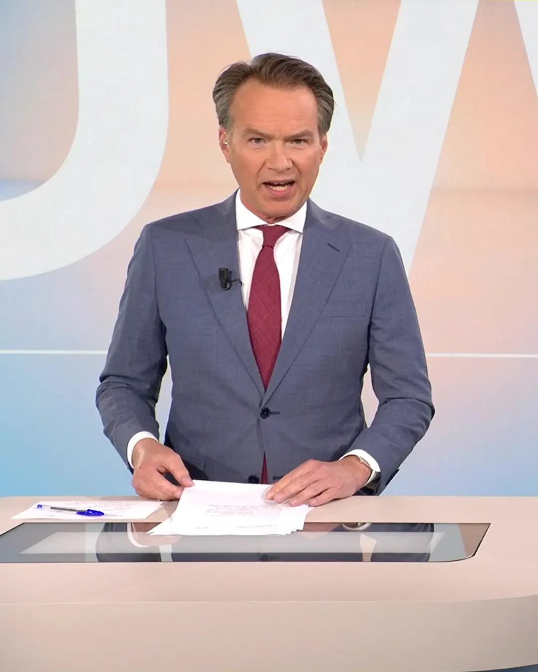 Male RTL Nieuws news anchor in blue suit and red tie presenting behind a desk with papers and a pen.