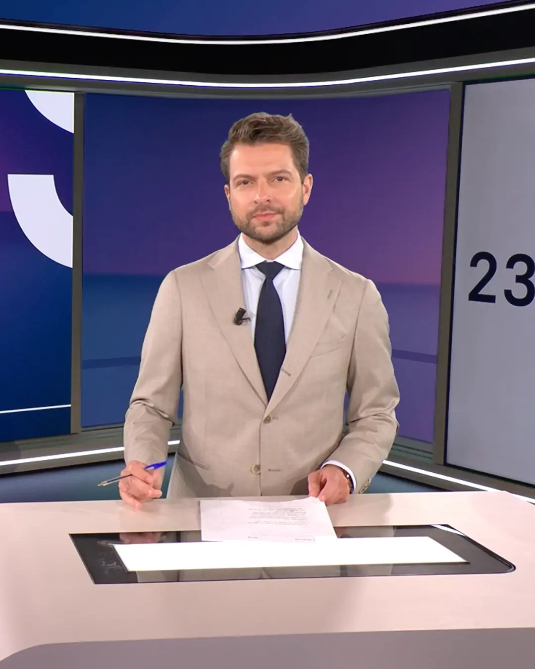 RTL Nieuws presenter in beige suit and navy tie standing behind news desk holding a pen and paper in a studio.