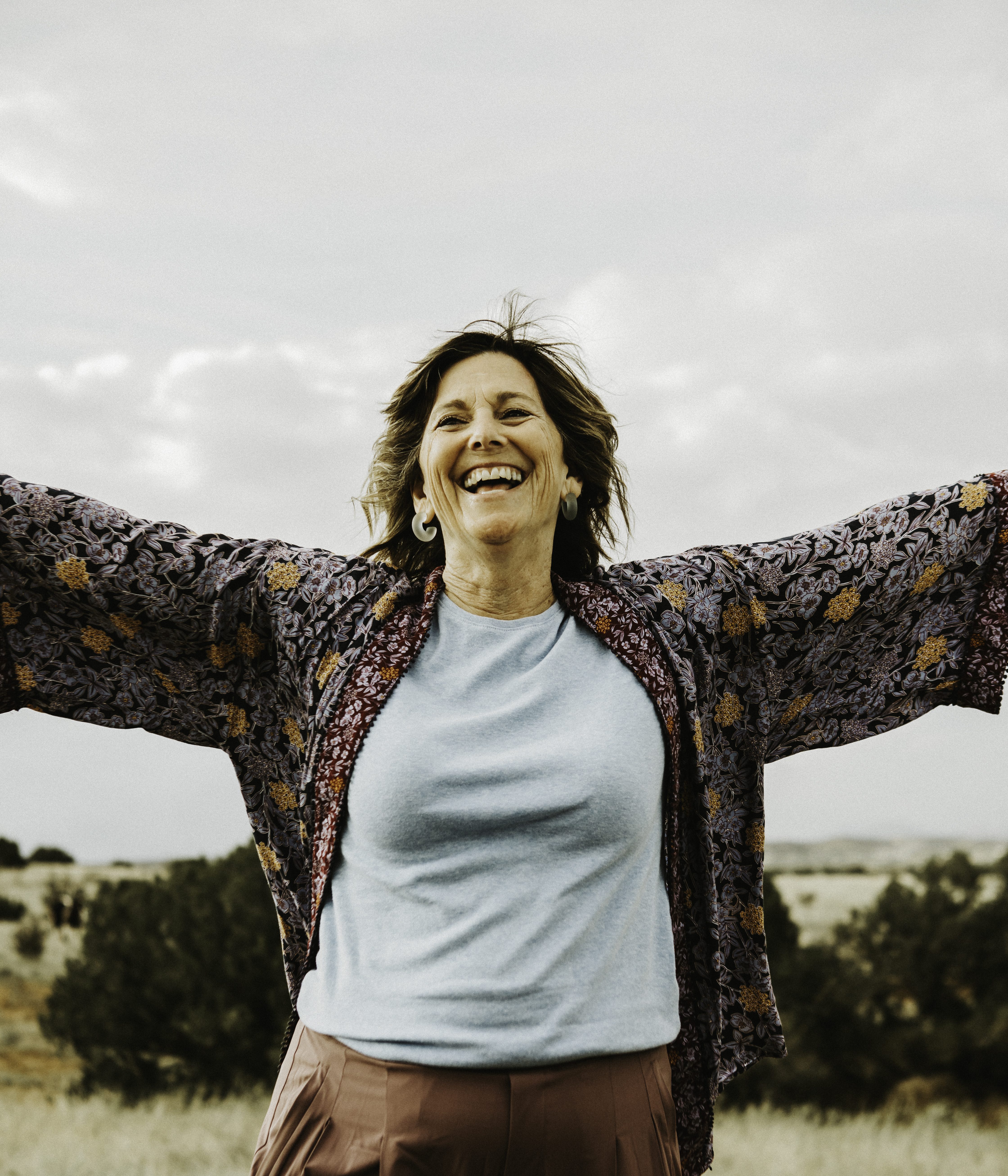 A smiling woman stands in front of junipers with her arms wide open