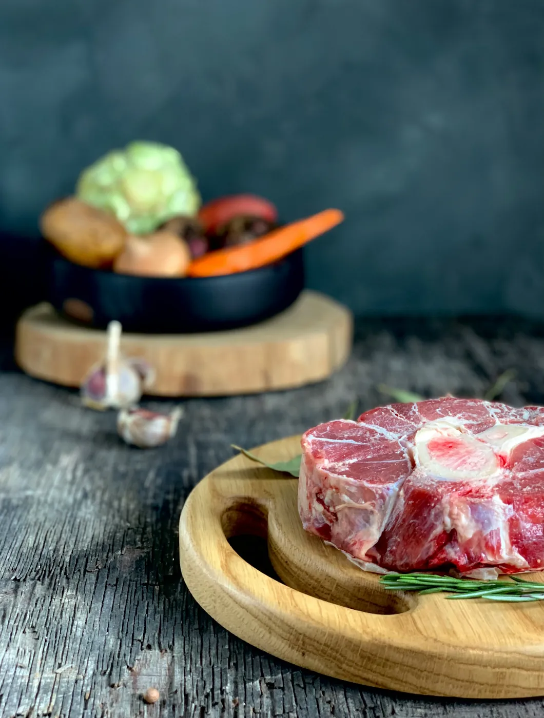 Une tranche de jarret de veau crue avec son os à moelle, sur une planche en bois, avec l'ail, le laurier et les légumes du bouillon