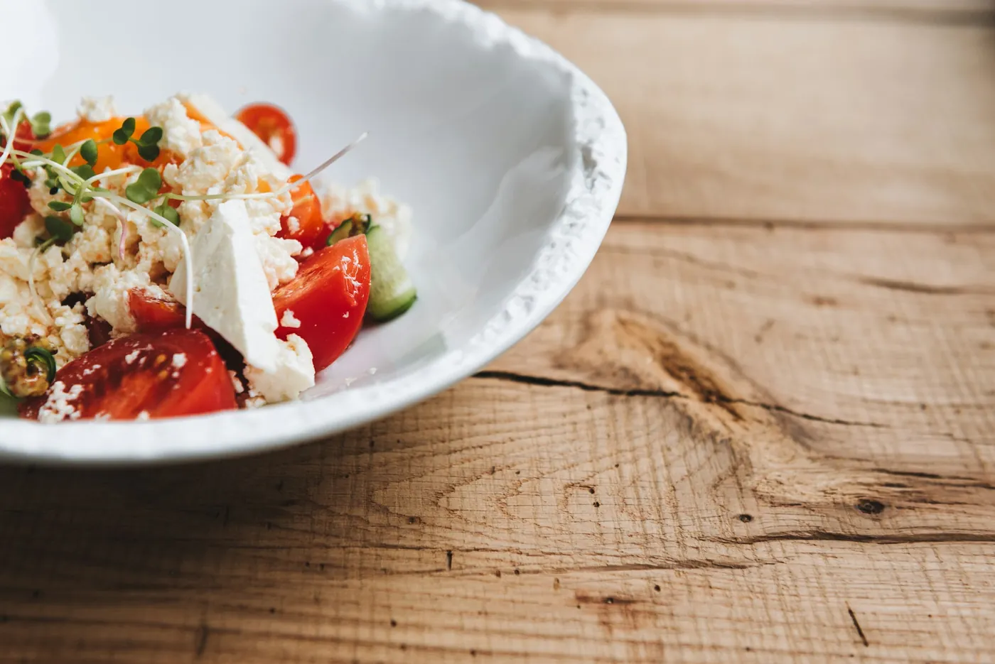 Une assiette creuse blanche garnie de quartiers de tomates, de feta émiettée et de rondelles de concombre, sur une table en bois brut