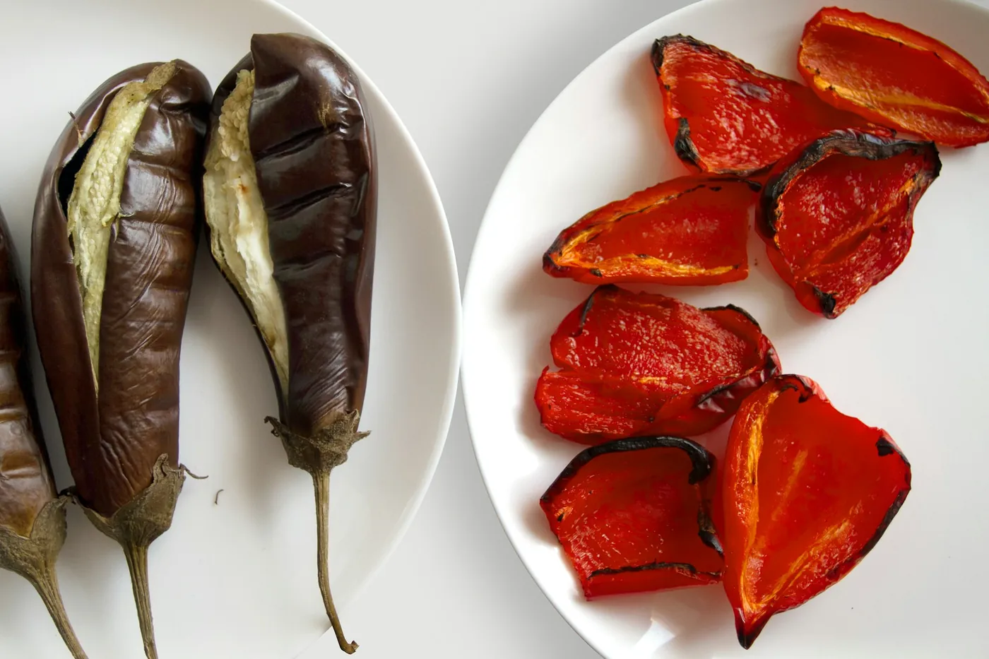 Deux assiettes blanches vues de dessus : des aubergines rôties fendues en deux et des lanières de poivron rouge aux bords noircis