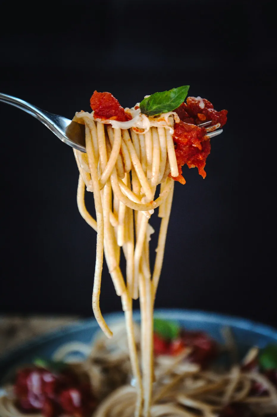 Une fourchette soulevant des spaghettis nappés de sauce tomate, avec une feuille de basilic, au-dessus d’une assiette