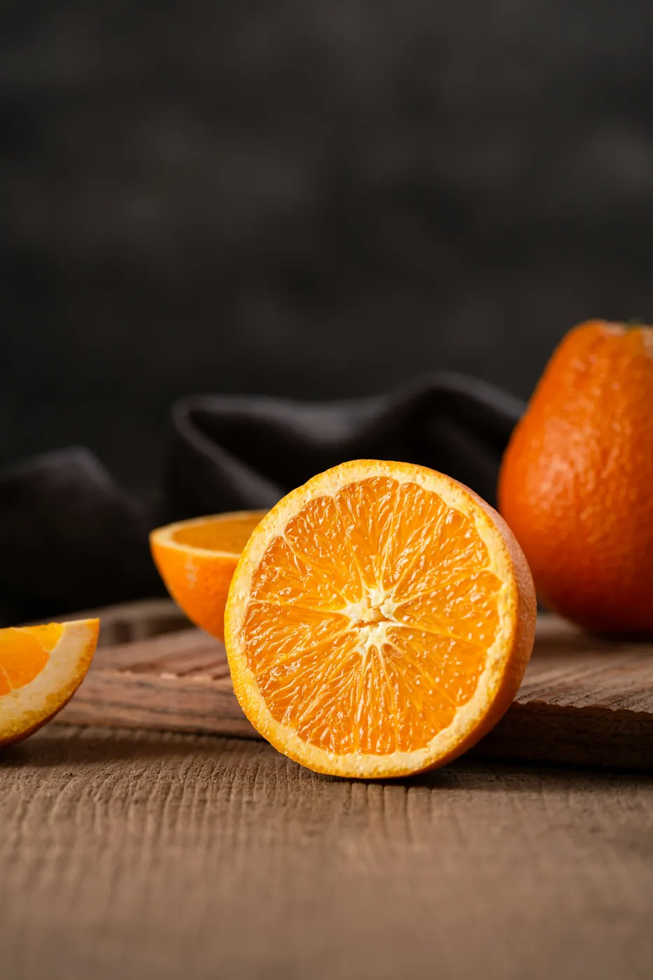 Une orange coupée en deux, un quartier et une orange entière posés sur une planche en bois, sur fond sombre