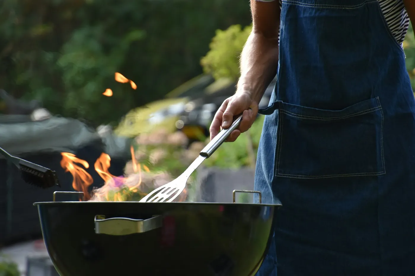 Une personne en tablier de jean attise un barbecue rond dans un jardin, spatule à la main, quelques flammes au-dessus de la grille