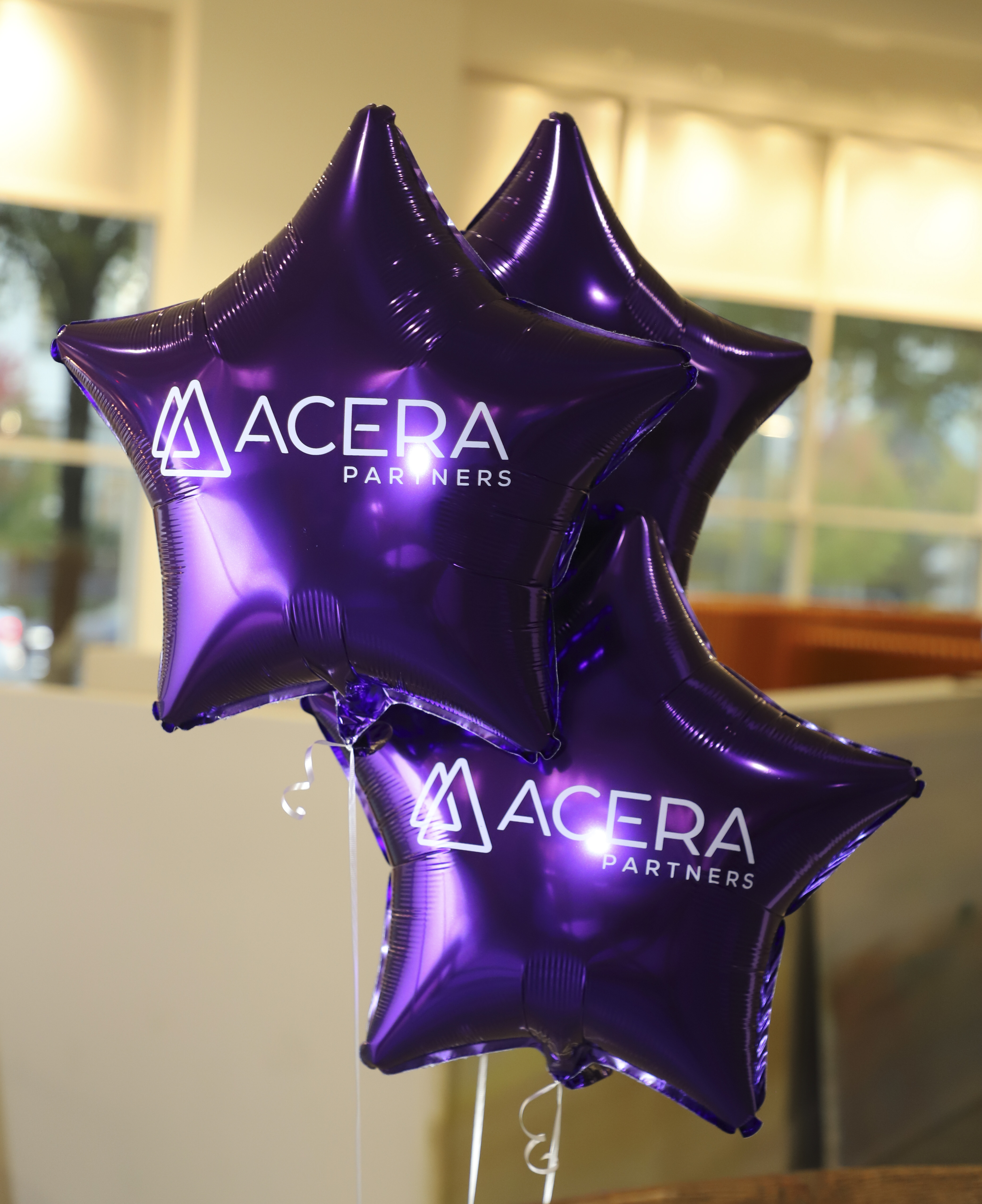 Three shiny purple star-shaped balloons with the white Acera Partners logo indoors.
