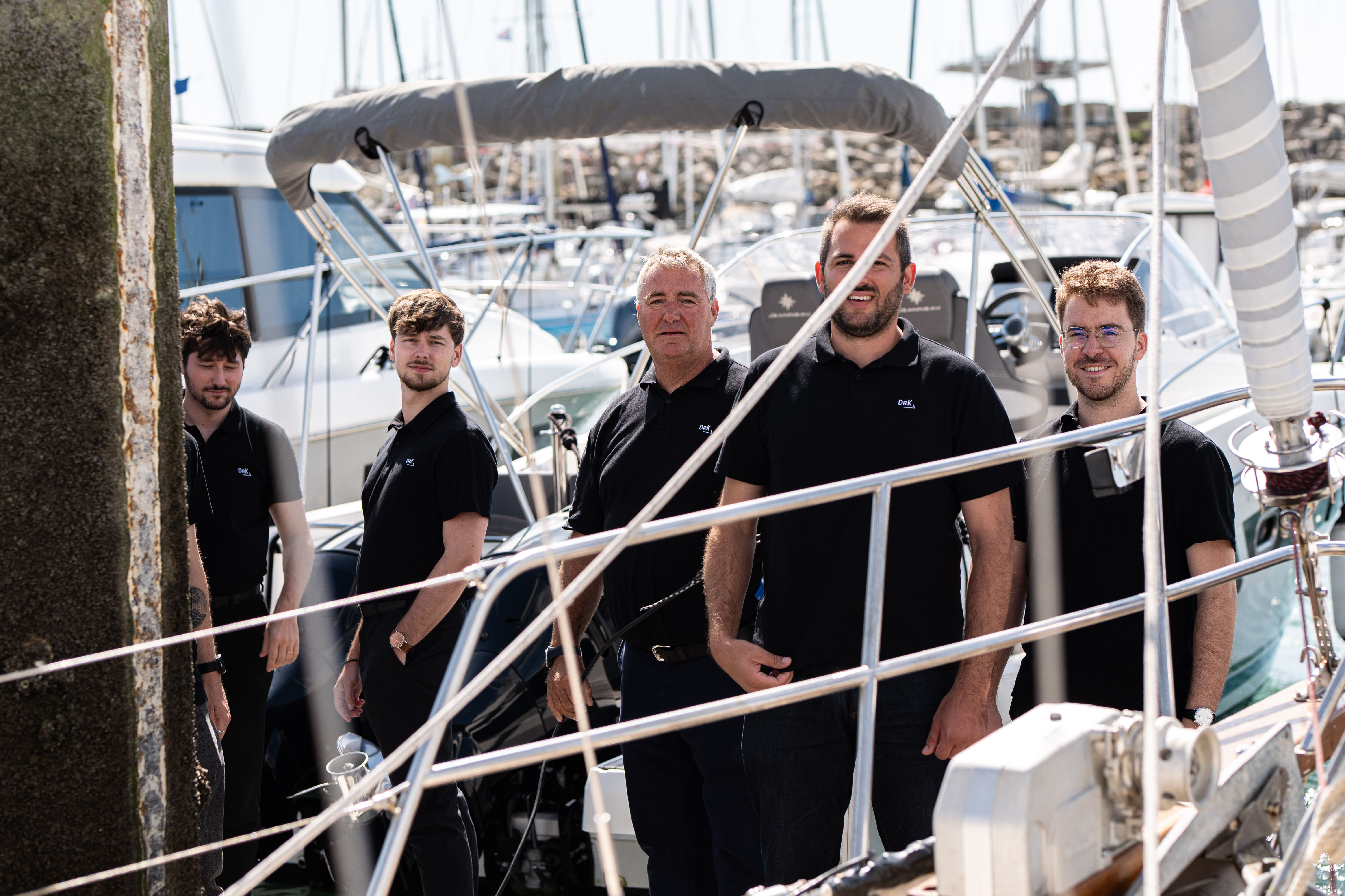 Cinq hommes en polos noirs posent sur un voilier dans un port avec d'autres bateaux en arrière-plan.