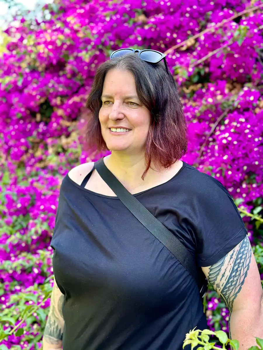 Frau in schwarzem T-Shirt mit Tätowierungen vor leuchtend violetten Blüten, trägt Sonnenbrille auf dem Kopf.