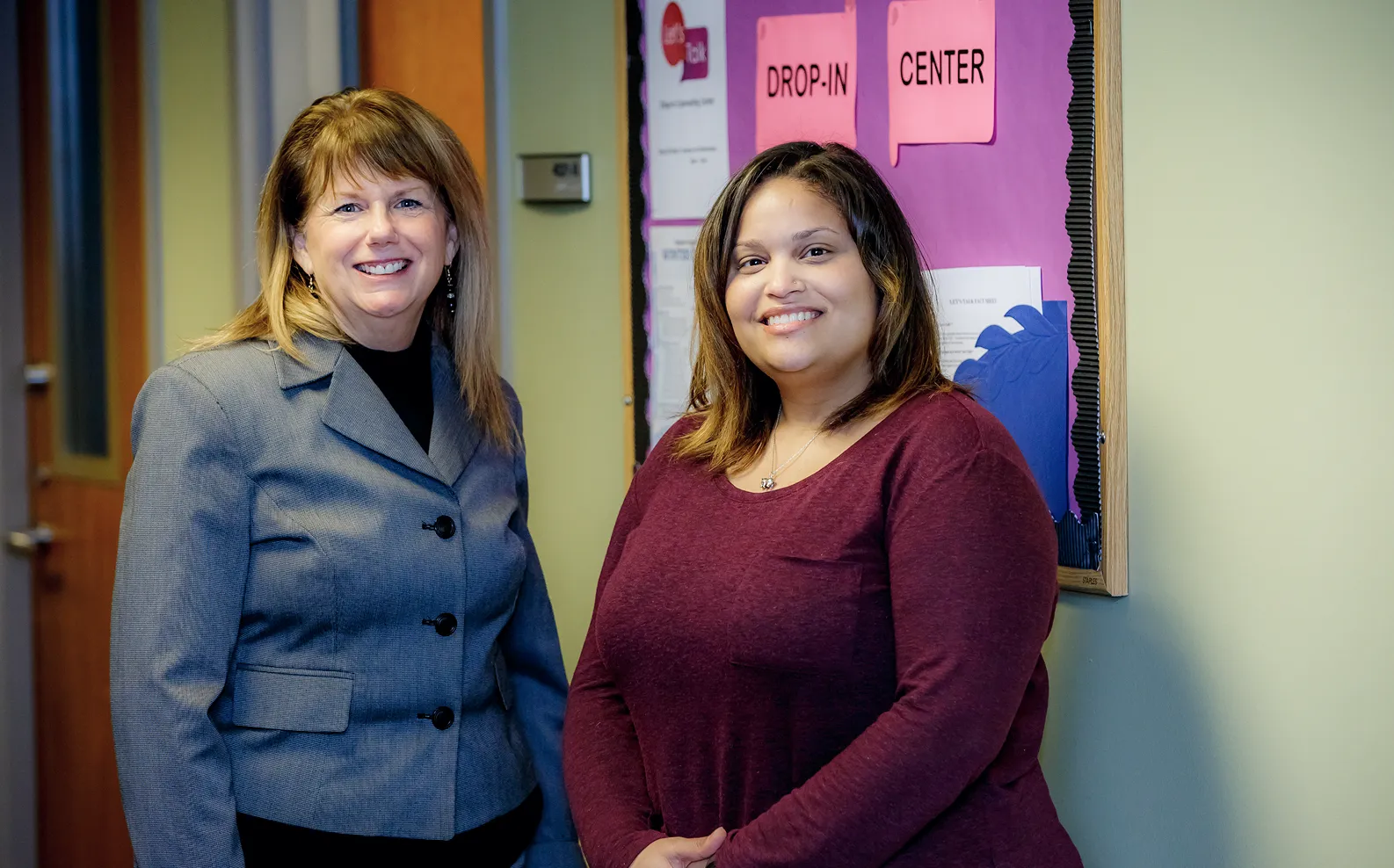 Two women standing next to each other in front of a bulletin board.