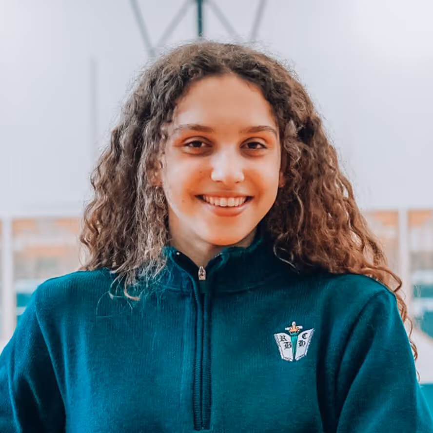 Smiling young woman with long curly hair wearing a green zip-up sweater indoors.