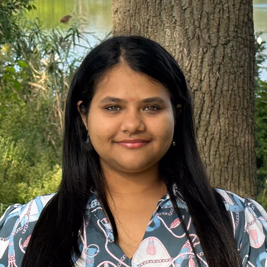Smiling woman with long black hair standing outdoors in front of a tree and greenery.