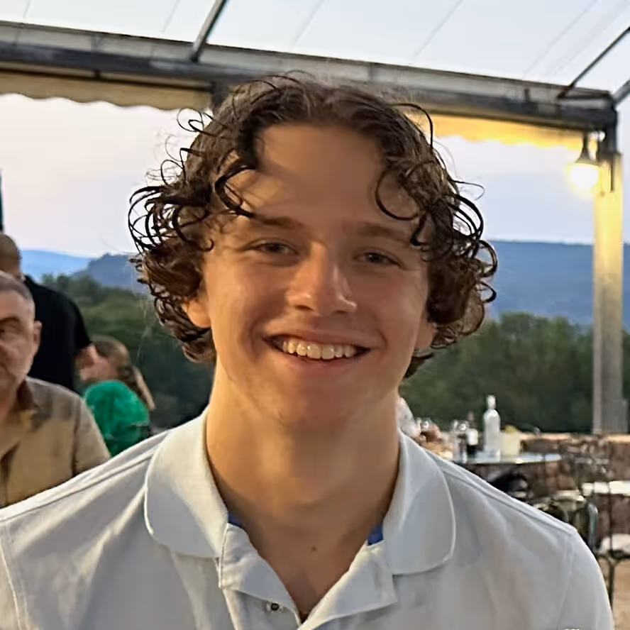 Young man with curly hair smiling, wearing a white collared shirt, seated outdoors under a canopy at dusk.