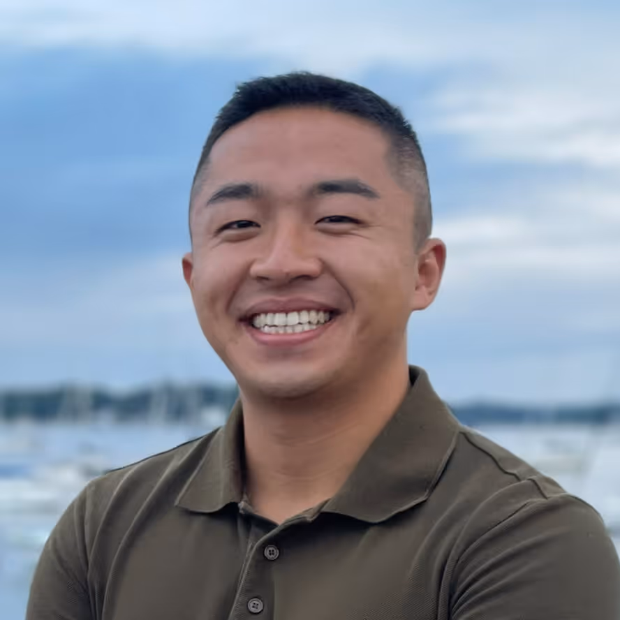 Smiling man in a dark green polo shirt standing outdoors with a blurred background of water and boats.