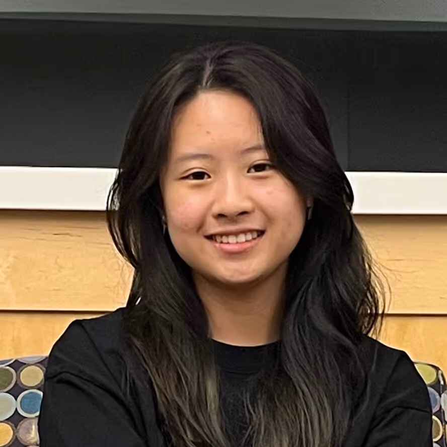 Young woman with long dark hair smiling indoors against a wooden panel background.