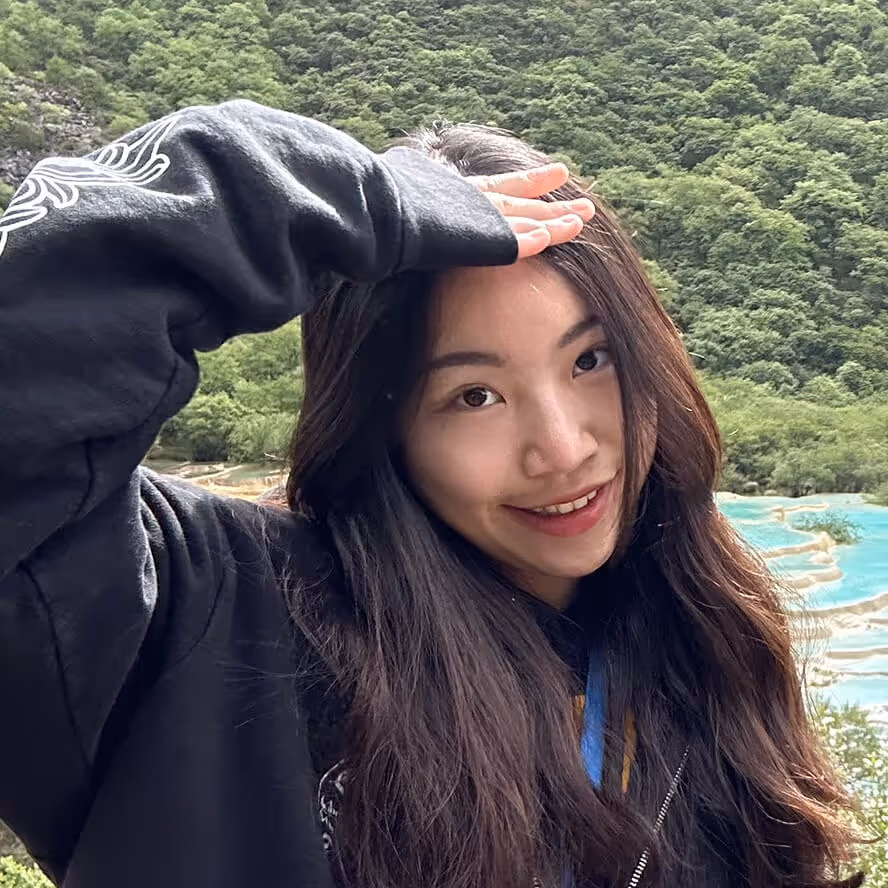 Young woman smiling with long hair, shielding her eyes from the sun with her hand, against a backdrop of green hills and blue water terraces.